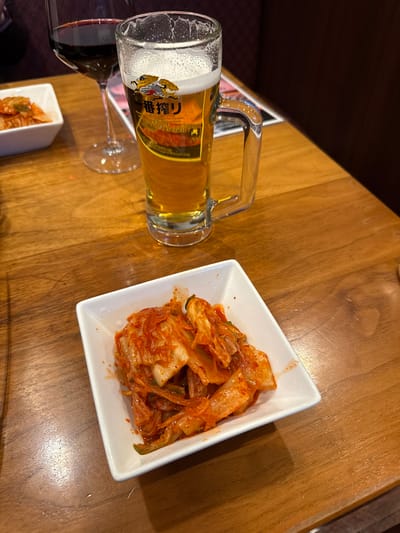 This image captures a tabletop scene inside a dining establishment in Nagano, Japan. The foreground features a white square dish containing a serving of reddish-orange kimchi. Immediately behind this dish, a clear glass beer mug, identified by its "一番搾り" (Ichiban Shibori) label, is filled with light amber beer topped with a frothy white head. To the left of the beer mug, a stemmed wine glass holds a dark red liquid. A second, partially visible white square dish with a similar food item is situated further back on the left. The table is constructed of polished wood. The background is blurred, showing dark, upholstered seating or wall panels, indicative of an indoor restaurant or bar environment. No people are visible.