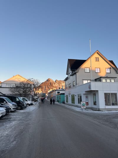 The image displays a street view in Henningsvær, Norway, under a clear, light blue sky with low-angle winter sunlight. The scene depicts a paved road, partially covered with snow and ice, extending from the foreground into the distance. On the left side of the road, a row of approximately five parked vehicles, predominantly dark-colored vans and cars, are situated on a snow-dusted shoulder.
To the right, a prominent two-story building with white siding and a dark pitched roof occupies the mid-ground. Its upper facade and windows are illuminated by warm, golden sunlight. A tall, white flagpole, devoid of a flag, is attached to its front. At street level, the building houses a commercial establishment with large glass windows, outside which a white sandwich board displaying red text, including "Tilbud -30%", and a red wall-mounted mailbox are visible.
Further along the street, other multi-story buildings with pitched roofs, painted in various light hues such as yellow, blue-green, and white, line both sides. Several individuals are present on the right side of the street: a group of three to four people walks together in the middle distance, followed by two more individuals. All are dressed in dark, heavy winter clothing, walking away from the viewer.
The background is dominated by a rugged, snow-dusted mountain peak, illuminated by direct sunlight, which gives its rocky face a warm, golden glow. Sparse, leafless trees are interspersed among the buildings on the left side of the street. Shadows are cast by buildings and objects, indicating the direction of the low sun.
