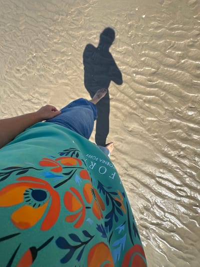 The image shows a person's legs and torso from a high-angle perspective, standing on wet sand at the beach in Berck, France. The person is wearing blue jeans and a teal-colored garment with a floral pattern of orange poppies. The shadow of the person is cast on the sand. The sand has ripples and is wet from receding water, indicating the proximity to the sea. The lighting suggests it is daytime, likely sunny.