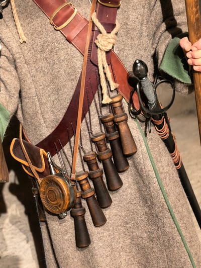 The image shows a close-up, waist-up view of a person dressed in historical clothing, likely a reenactor, in Staraya Ladoga, Russia. The individual is wearing a rough, tan woolen coat with green fabric visible at the cuffs. A wide leather belt, secured by a brass buckle, is worn over the coat. Suspended from the belt are several items: a leather pouch, a small round flask made of wood and metal, and a series of wooden powder horns. A sword with a dark hilt and metal guard is sheathed in a black scabbard adorned with leather straps. A wooden staff or spear is visible on the right side of the frame, held by a hand. The background is a blurred, indoor setting.