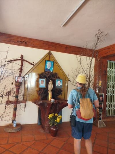 The image depicts a person wearing a light blue t-shirt, dark blue shorts, a yellow hat, and a tan backpack. The person is standing in a room with terracotta-colored floor tiles. In front of the person is a makeshift altar against a light-colored wall with a wooden beam running horizontally near the ceiling. The altar features three framed portraits of men, a statue of the Virgin Mary, and a wooden cross. To the left of the altar is a crude wooden cross-like structure. To the right of the altar is a tall, leafless tree branch. A vase with yellow flowers sits at the base of the altar. A barred gate is visible in the background to the right. The location is identified as An Phu Ward, Vietnam.