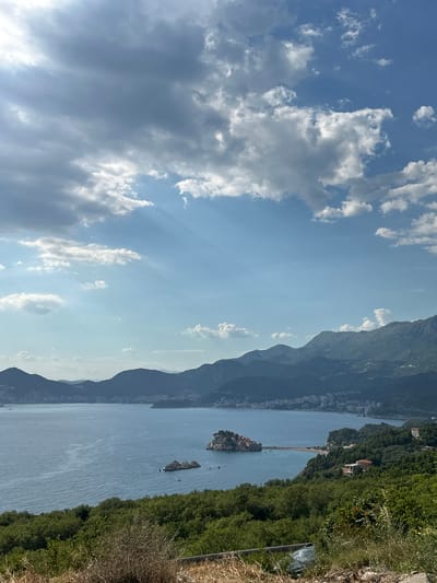 This is a vertical, eye-level, medium shot of the coastal region near Blizikuće, Montenegro, on a day with a partially cloudy sky and sunbeams breaking through. The foreground is dominated by dry, brown vegetation and some green foliage. In the mid-ground, a lush green hillside slopes down towards the Adriatic Sea. The sea itself is a deep blue, with a few small boats visible. Dominating the mid-ground is the distinctive island of Sveti Stefan, a small fortified village connected to the mainland by a narrow causeway, with its reddish-brown rooftops and surrounding green vegetation. Beyond the island, the coastline curves, revealing more green hills dotted with buildings and a distant city or town. The background consists of a range of layered, dark blue mountains, with the sky above featuring both bright blue patches and dense, gray clouds.