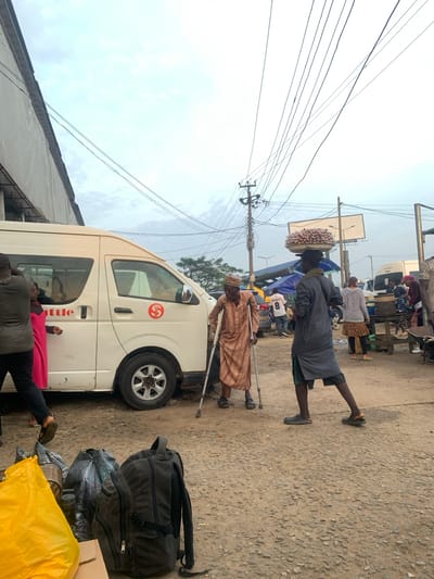 In Elelenwo, Nigeria, an outdoor scene unfolds with a white shuttle van on the left. A man in a brown, patterned robe and a cap, using two crutches for support, stands near the van. To his right, another man dressed in a dark gray robe and shorts carries a large tray of food items on his head. Behind them, several other individuals are visible, along with market stalls and various vehicles, suggesting a busy market or transportation hub. The ground is unpaved and covered with gravel and dirt, typical of the area. Power lines crisscross the sky above. In the foreground, several bags, including a yellow plastic bag and a dark backpack, are placed on the ground.