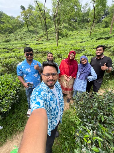 The image shows a group of six individuals posing for a selfie in a lush, green tea garden. The location is identified as Sylhet, Bangladesh, known for its extensive tea plantations.
In the foreground, the arm of the person taking the selfie is visible, wearing a blue and white patterned shirt. To their right, a man wearing glasses and a similar blue and white shirt is smiling. Behind them, from left to right, are: a man in a blue shirt with a cloud pattern, giving a thumbs-up; a man in a black polo shirt, also giving a thumbs-up; a woman wearing a red hijab and a cream and red patterned outfit; and a woman in a blue hijab and a light blue patterned top. They are all standing amongst rows of tea plants that cover rolling hills. The environment is outdoors with trees scattered on the slopes of the tea garden, and the sky appears overcast. The overall scene suggests a casual outing or visit to a tourist attraction in Sylhet.