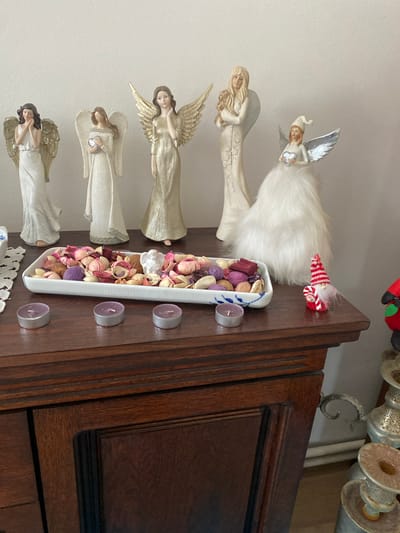 A still life arrangement is displayed on a dark wooden cabinet. In the background, five angel figurines stand against a white wall, arranged from left to right: a blonde angel with a child, a blonde angel holding a heart, a blonde angel with golden wings, a dark-haired angel with golden wings, and a dark-haired angel in a white dress with golden wings. In the foreground, a white rectangular dish filled with potpourri sits on the cabinet, accompanied by a small white angel figurine. Three small, unlit purple tea lights are placed in front of the dish. To the right of the dish, a small gnome figurine in a red and white striped hat, holding a candy cane, is positioned next to a red and green ornament. The cabinet itself is dark brown wood with visible grain and decorative molding. The overall scene is well-lit, with the light source appearing to be from the right, casting subtle shadows. The location is not specified in the image, but if it were in Osek, Czechia, it would be an indoor domestic setting.