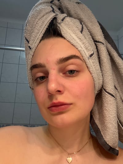 A young woman with a towel wrapped around her head poses for a close-up photograph in a bathroom. She has light skin, blue-grey eyes, and full lips. A gold necklace with a heart-shaped pendant is visible around her neck. The bathroom features tiled walls, with tiles visible in the background. The location cannot be determined definitively as Hainburg an der Donau, Austria from the image.