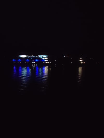 At night, the photo shows a body of water with a ship in the distance. The ship is illuminated with white and blue lights, and the lights are reflected in the water. There are some buildings in the distance with lit windows. The water is dark and still. The photo appears to be taken at night in Tikhonovo, Russia.
