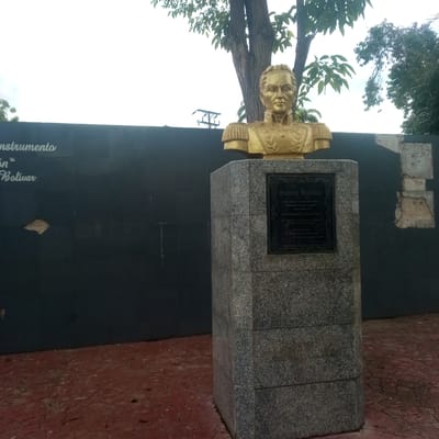 The image captures a bronze bust of Simón Bolívar, a prominent historical figure, displayed on a granite pedestal in what appears to be a public space in El Limón, Venezuela. The bust, rendered in a gleaming gold-bronze hue, depicts Bolívar in military attire, featuring epaulets and a high-collared jacket adorned with laurel wreaths. A dark plaque with ornate borders is affixed to the front of the pedestal, bearing text in a decorative font that likely details Bolívar's name and a significant date, possibly his birth and death years, "1783 - 1830". The pedestal itself is constructed from grey granite tiles.
Behind the bust, a dark, textured wall serves as a backdrop, possibly a mural or a painted surface. Faint white lettering is visible on this wall, hinting at an inscription or artwork related to "instrumento" and "Bolívar". To the left of the wall, the white expanse of the sky is partially obscured by the lush green foliage of trees. To the right of the pedestal, the wall shows signs of wear, with sections appearing to be chipped or broken, revealing lighter material underneath.
The ground in the foreground is paved with red brick or tiles, giving the impression of a plaza or walkway. The overall scene is set outdoors, under what seems to be an overcast sky, with natural light illuminating the bust. The environment suggests a place of historical significance or commemoration within the city of El Limón, Venezuela.