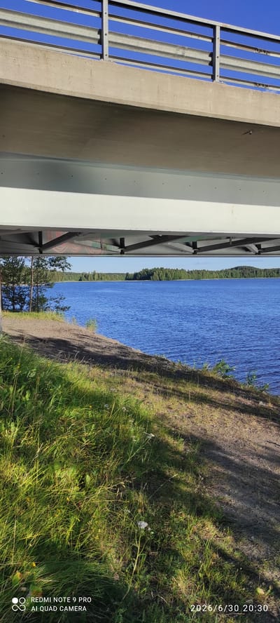 The image depicts a tranquil scene of a body of water, likely a lake, with a dense forest lining its distant shore. The water is a vibrant blue, reflecting the clear sky above. In the foreground, a grassy bank slopes down to the water's edge, dotted with wildflowers and long, green grass. The sun is casting long shadows across the terrain, suggesting it is late afternoon or early evening.
On the left side of the image, a portion of a concrete structure, possibly a bridge or a building, is visible. It features metallic railings and vertical supports, suggesting an urban or infrastructure element intersecting with the natural landscape.
Visible text includes a date and time stamp: "2026/6/30 20:30" and "REDMI NOTE 9 PRO AI QUAD CAMERA". This information indicates when and with what device the photo was taken. The overall mood of the image is serene and peaceful, capturing a moment of natural beauty in Finland, specifically Hartola. The weather appears to be pleasant, with clear skies and ample sunlight.