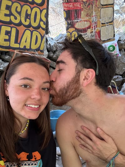 A close-up image depicts two young adults in an outdoor setting in San Miguel Atlapulco, Mexico. On the left, a female with medium-brown hair, light brown eyes, and a light complexion smiles directly at the camera. She wears a black top with an orange and yellow design, a yellow beaded necklace, and small silver hoop earrings. Light-colored sunglasses are perched on her head. To her right, a male with dark hair and a reddish-brown beard and stubble is shown with his eyes closed, kissing the female's left cheek. He appears shirtless, with dark sunglasses resting on his head, and a multi-colored braided bracelet visible on his left wrist. His left hand rests flat against his upper chest. The background features a textured stone wall or rock formation. Several weathered commercial signs and posters are affixed to the wall, including a yellow sign with blue lettering partially legible as "ESCOS ELADA" and other posters featuring "Coca-Cola" and "Modelo" branding. A light blue bucket, partially filled with water, is visible in the mid-ground behind the male's arm, and a white bag is visible in the upper right background.