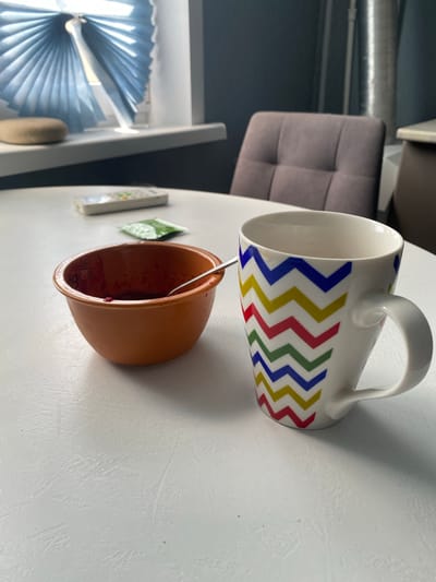 This is a close-up, eye-level shot of a breakfast setting on a textured white table. To the left, a small, orange ceramic bowl contains a dark red substance, likely jam or compote, with a silver spoon resting inside. Adjacent to the bowl is a small green packet, possibly sugar or sweetener, and a white remote control. To the right of the bowl is a white ceramic mug with a colorful chevron pattern in blue, yellow, red, and green. The mug is empty and shows a slight stain near the rim. In the background, a gray upholstered chair with a tufted design is visible, along with a partially obscured kitchen counter. A window with a blue pleated blind is also present, casting diffused light. The scene appears to be indoors, likely in a kitchen or dining area. The overall mood is quiet and domestic. The image is undated and does not provide any specific information about its location within the city of Sakmara, Russia.