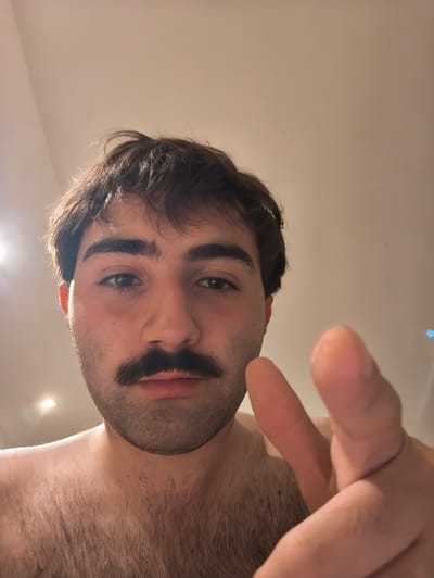 A young man with short brown hair, a mustache, and a bare chest is pointing his index finger at the camera. The man's face is in focus, and his torso and arm are out of focus. The background appears to be a plain white wall. This image was taken in Saint-Josse-ten-Noode, Belgium. The photo's location is not discernible from the image. There are no other people or objects in the image.