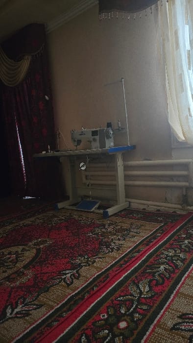 A sewing machine stands on a blue and white metal table, next to wall-mounted pipes. The machine is gray and has an oil gauge visible. The floor is covered with a red and tan patterned rug. Part of a curtain is seen hanging from the wall at the left.