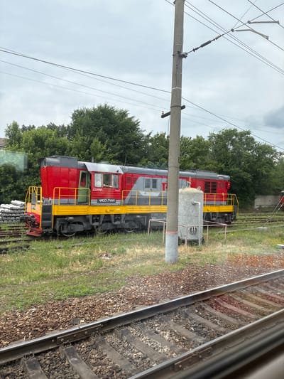 The image shows a red and gray diesel locomotive with yellow accents parked on a railway track. The locomotive has Cyrillic lettering on its side, including "ЧМЭ3-7444". A person can be seen through the locomotive's window, with their arm resting on the sill. In the background, there are trees, a utility pole with overhead wires, and a large cylindrical metal tank. The scene appears to be in a railyard or industrial area, with additional railway tracks and ballast visible in the foreground. The sky is overcast. The provided text in the image is in Russian. The location of Povorino, Russia, is mentioned in the prompt.