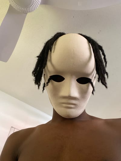 An individual with a dark skin tone is depicted from the upper chest upward, standing in what appears to be an indoor environment within Capesterre-Belle-Eau, France. The person is wearing an expressionless, smooth, cream-colored mask that covers the entire face, featuring oval-shaped, empty eyeholes. Dark, matted hair, consistent with dreadlocks, emerges from the sides and top of the mask, framing the upper portion of the head. The individual is shirtless, exposing bare shoulders and upper chest. The background consists of a light-colored, possibly off-white, wall and ceiling. A white ceiling fan with multiple blades is partially visible in the upper left quadrant of the image. No other individuals or significant objects are present, and no specific actions or interactions are discernible beyond the subject posing for the photograph.
