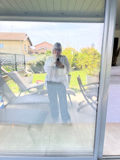 The image displays a layered view, captured through a reflective, dust-speckled glass door or window, in Saint-Just-Saint-Rambert, France, during daylight hours. A woman with light-colored hair, wearing a white long-sleeved top and dark trousers, is visible in reflection, holding a smartphone with both hands, suggesting she is taking the photograph.

Her reflection overlays an outdoor garden scene visible through the glass. This garden features dense green and reddish foliage, a grey striped lounge chair, and other outdoor furniture including a dark garden chair and a glass-topped table. Several potted plants are positioned on a paved patio area. In the background, two distinct residential houses are discernible: one light yellow with a terracotta roof and an antenna, and another light pink. A triangular white sign with a red border is partially visible through the distant foliage. The sky above is bright with light cloud cover. The interior space from which the photograph was taken is subtly reflected, revealing a grey upholstered furniture piece and a tiled floor. The glass surface itself shows numerous small specks, indicative of dust or minor smudges.