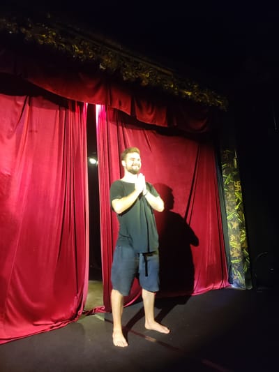 A medium shot captures a barefoot man with a beard, wearing a black t-shirt and blue shorts, standing on a stage. His hands are together in a prayer-like gesture. Behind him are red stage curtains, and to the right is a vertical column with a patterned covering. The stage appears to be in Bedulu, Indonesia.