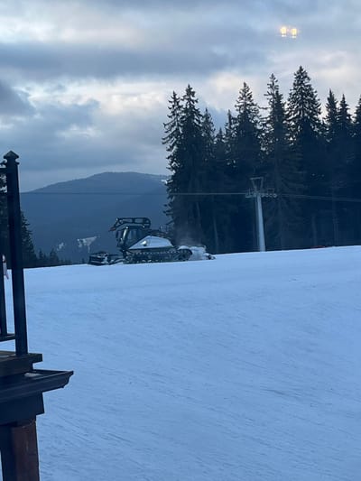 The image depicts a snow-covered ski slope in Stoikite, Bulgaria, under an overcast sky, likely during dusk or dawn. In the midground, a large, silver-grey and black snow groomer with tracks is actively moving rightward across the slope, pushing snow, indicated by disturbed snow behind its rear and a faint plume. No operator is visible within its cabin. In the immediate foreground, to the lower left, a dark wooden support post and a section of a dark, horizontal structure, possibly a railing or roof edge, partially obscure the view. The background features a dense, dark evergreen forest of conifers and a grey ski lift pole positioned within the trees to the right of the groomer. Farther in the background, dark, tree-covered mountain ranges are visible beneath a mostly grey, cloudy sky. Two distinct, bright artificial lights are visible high in the upper right sky, suggesting a light source out of frame. The environment is marked by extensive snow cover typical of winter.