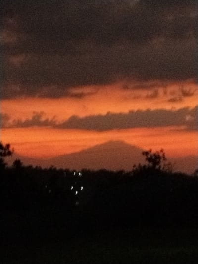 The image depicts a dark, grainy silhouette of a conical mountain against a vibrant sunset sky. The sky is a gradient of deep oranges and reds, with darker clouds forming horizontal bands across it. A dark, jagged treeline forms the foreground, with a few blurry, white lights visible amongst the trees, possibly indicating distant streetlights or dwellings. The overall scene suggests a rural or semi-rural setting at dusk, with the imposing silhouette of the mountain dominating the background. The location in Menjing, Indonesia, is not explicitly visible in the image but would be the geographical context of this natural landscape.
