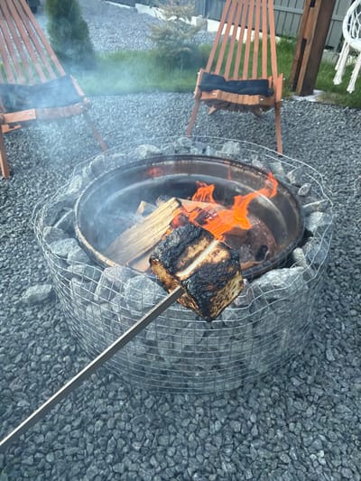 In the city of Sharovechka, Ukraine, a fire pit constructed from rocks and wire mesh contains burning wood and flames. A metal skewer holds a large, charred marshmallow over the fire. Two wooden chairs with black cushions are positioned to the left and right of the fire pit, with one slightly angled towards the camera. The ground surrounding the fire pit is covered in gray gravel. In the background, a green bush is visible to the left, and a wooden fence and a white macrame chair are present on the right. Smoke is gently rising from the fire pit.