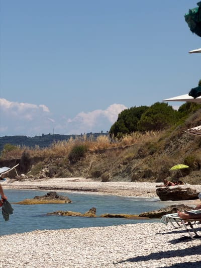 This is a landscape photograph taken in the Elaties Municipal Unit, Greece. The image depicts a rocky beach with clear turquoise water. The beach is covered in small white pebbles, and the water is shallow near the shore, becoming deeper further out. The scene is bordered by a steep, vegetated hillside. The hillside is covered in dry grasses and some scattered green shrubs and trees, suggesting a warm, dry climate. 

In the upper right of the image, a person is seated on a beach chair, with their feet visible. They are partially shaded by a small, green umbrella. Further along the beach, another person is visible, also seated. In the foreground, a person's hand is holding a dark green fabric. 

The sky is a clear, bright blue with a few scattered white clouds, indicating it is daytime, likely a sunny day. The lighting suggests it is mid-morning or late afternoon, given the long shadows cast by the vegetation and beach chairs on the pebbles. The overall mood of the image is peaceful and relaxing, evoking a sense of a summer vacation. There is no visible text in the image.
