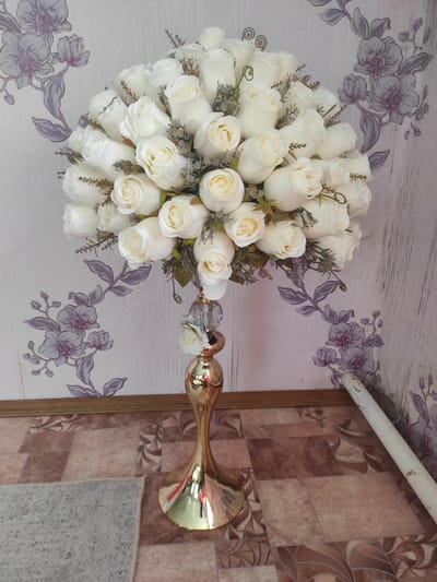 The image shows a close-up of a floral arrangement of white roses in a gold vase. The vase has a decorative, twisted stem and sits on a floor with a tiled pattern in shades of brown and beige. The background features wallpaper with a floral design in shades of purple and gray. A section of a white pipe is visible on the right side of the image, leaning against the wall. The scene appears to be indoors. No specific location in the city of Некрасовка, Kazakhstan can be determined from the visual information provided.