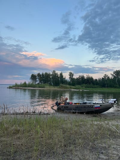 A person in camouflage clothing is partially submerged in water, leaning over the side of a small motorboat docked on a sandy shore. The boat has the registration "AB2301RUS54" visible. The scene is set at dusk, with a body of water in the foreground and a treeline along the distant shore. The sky is a gradient of soft blue, pink, and gray clouds, reflecting on the calm water. The location appears to be a natural landscape, possibly a lake or river, with tall grasses growing along the shoreline. The general atmosphere is tranquil and serene, indicative of a quiet evening by the water.
