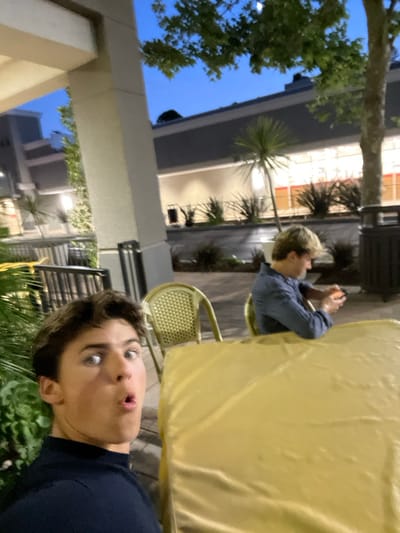 The image depicts an outdoor commercial setting at dusk or early evening, located in Rolling Hills Estates, United States. In the immediate foreground, a young male with light brown hair and blue eyes, wearing a dark collared shirt, is shown from the chest up, facing the camera with an open-mouthed, wide-eyed expression. A large, rectangular table draped in a smooth, golden-yellow fabric occupies the lower right portion of the frame. To the right of this table, another young male with blonde hair is seated in a light-colored wicker chair, viewed from the chest up. He wears a blue long-sleeved shirt and is focused on a small, dark electronic device held in both hands. An empty matching wicker chair is visible to his left. The background features a multi-story commercial building with light-colored walls, a dark roof, and numerous brightly lit windows revealing interior spaces, possibly a retail establishment. The building is fronted by landscaped areas containing various shrubs and small palm-like trees. The sky is a deep twilight blue, and several streetlights or building lights are illuminated. Architectural columns and dark railings of the immediate structure are visible in the left and lower-left portions of the image.