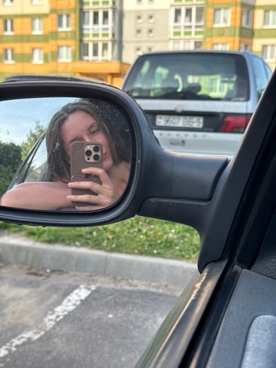 In the city of Zhodzina, Belarus, a young woman with braided hair takes a selfie in a car's side mirror. She is leaning on her left arm, with her right hand holding a smartphone in a brown case, angled to capture her reflection. The background of the mirror shows trees and a bright sky. The exterior of the car, a light-colored SUV with a license plate reading "9607 BE-5," is visible behind the mirror. The surrounding environment includes a curb, asphalt, and a grassy area. In the further background, there is a multi-story building with yellow and green facade elements. The image has a grainy texture, suggesting it was taken with a mobile phone in natural light.