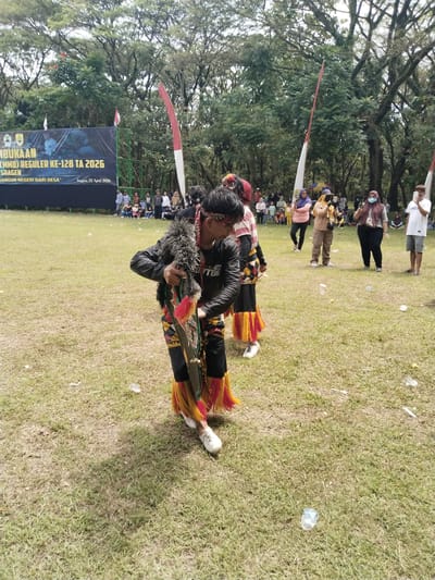 This image captures an outdoor cultural or official event taking place on a sunny day in a grassy field, likely in Puro, Indonesia.

In the foreground, a person dressed in elaborate traditional attire is prominent, possibly a dancer or performer. They wear a black jacket over a patterned top, black pants with vibrant red and yellow fringed trim, and white shoes. A decorative headband adorns their head, and they hold a long, ornate, multi-colored prop or instrument. Another person in similar traditional clothing is partially visible behind them.

The midground features an audience of spectators standing and watching the event. Several individuals are seen holding smartphones, seemingly recording or photographing the performance. Among them, a woman in a brown hijab and another in a yellow hijab holding a camera are noticeable. Red and white flags, resembling the Indonesian national flag, are positioned on poles, indicating a national or official ceremony.

In the background, a large black banner is erected, bearing white text. The legible text includes "PEMBUKAAN (TMMD) REGULER KE-128 TA 2026 SRAGEN BANGUN NEGERI DARI DESA" and "Sragen, 22 April 2026". This text suggests an opening ceremony for a community development program (TMMD) in Sragen. Behind the banner and the crowd, a dense array of tall, leafy trees forms the backdrop, giving the setting a natural, open-air feel. The ground is a mix of green and drier grass, with some discarded plastic litter visible. The scene conveys a lively community gathering focused on cultural performance and a formal inauguration.