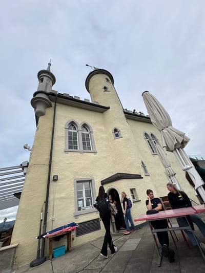 The image displays an outdoor patio area adjacent to a pale yellow, castle-themed building with grey architectural details under an overcast sky in Dulliken, Switzerland. The building features two distinct towers: a smaller, slender cylindrical tower with a pointed grey spire on the left, and a taller, broader cylindrical tower with a rounded grey cap and flag element on the right. The exterior walls are textured stucco, adorned with grey stone window surrounds, including two prominent arched windows on the upper floor and a rectangular window on the lower floor. A recessed, dark-roofed entrance is centrally located on the lower facade.

In the foreground right, two individuals, appearing to be young adults, are seated at a red rectangular outdoor table with grey metal legs. A male in a dark t-shirt is seated on the left side of the table, facing right and engaged in conversation with a female on the right side. The female has blonde hair and wears a dark long-sleeved top with yellow text. Two large, folded, light-colored patio umbrellas are positioned vertically next to the building on the far right.

Near the building's entrance, three additional individuals are present. One female, wearing a black long-sleeved top and black wide-leg pants, carries a black backpack and walks away from the entrance towards the left of the frame. Another female, with blonde hair, stands to the right of the entrance wearing a dark polo shirt, blue jeans, and a purple backpack. A third individual is partially obscured within the entrance. Small, indistinct figures are visible on the building's roofline near the base of the taller tower and behind one of the folded umbrellas.

On the lower left, a rustic wooden stand holds a red and blue striped umbrella and a turquoise plastic container. The ground is paved with light grey concrete or stone pavers. A modern, horizontally-slatted metal structure is visible in the distant upper-left background, suggesting an elevated vantage point overlooking distant hills.