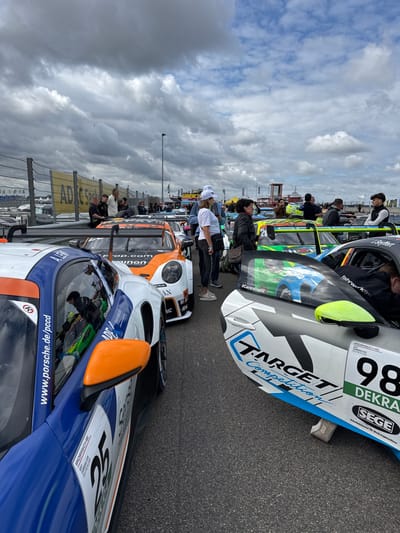 The image depicts a race event paddock or grid at the Sachsenring circuit in Hohenstein-Ernstthal, Germany. Under an overcast sky, multiple race cars are staged on an asphalt surface. In the immediate foreground left, a white and blue Porsche 911 GT3 Cup car, number 25, with an orange side mirror and "www.porsche.de/pccd" branding, is prominently displayed. Adjacent to it, an orange and white Porsche 911 GT3 Cup car is positioned. On the immediate foreground right, a white race car with black and light blue "TARGET Competition" graphics and number 98 is visible. Numerous other race cars, primarily Porsche 911 GT3 Cup models, are discernible in the mid-ground. Approximately 15 to 20 individuals, dressed in casual attire and some in team apparel, are scattered throughout the scene, standing, observing the vehicles, and conversing. A chain-link fence, topped with a yellow banner displaying "ADAC Sachsen", extends across the background, behind which additional vehicles and a red tower structure are visible. The overall environment indicates a live motorsport event.