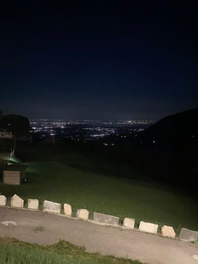 The image depicts a nighttime view of a city from a high vantage point, likely a hillside overlooking Caprino Veronese, Italy. The foreground shows a gently sloping grassy area with a path and a low border of light-colored stones. Further down the slope, a small wooden structure is visible. In the middle ground, a tree casts a dark silhouette against the distant city lights. The background is dominated by a sprawling urban landscape illuminated by countless lights, suggesting a significant population center. The sky above is dark, with a faint hint of stars or distant celestial bodies visible. The overall impression is one of a peaceful, illuminated city viewed from a quiet, natural setting at night.