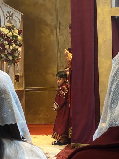 A vertical, medium shot depicts a scene within a church, possibly located in Södertälje, Sweden, based on architectural and religious cues. A young man in a red, embroidered robe and black shoes is the central subject. He appears to be bowing, possibly in reverence or during a religious ceremony. Another person, possibly a priest or religious figure, is partially visible near the top left, wearing an ornate golden garment, with their hand extended.

The environment is characterized by a painted gold wall. In the foreground, white tulle is partially obscuring the view. Below and to the left, a floral arrangement consisting of white and red roses sits atop an ornate structure with a cross. Red drapery hangs above the subjects.