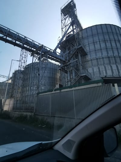 The image shows a series of metallic grain silos located in Razdelna, Bulgaria. The silos are connected by a network of metal conveyor belts and supporting structures. A corrugated metal fence runs along the base of the silos. The photograph was taken from inside a vehicle, with the dashboard and side mirror visible in the foreground. The sky is light blue.