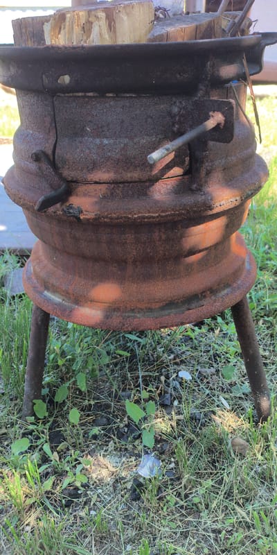 A close-up, vertical, outdoor, eye-level shot shows a rustic, rusty metal stove with a wide, flared opening at the top, filled with chopped wooden logs. The stove is cylindrical with a ridged, layered design and sits on three metal legs. A small, rectangular door with a simple metal latch is visible on the front of the stove. The metal is heavily oxidized, with patches of orange and brown rust. The stove is situated on a patch of dry grass and dirt, with some green weeds growing around it. The background is blurred, suggesting a natural or rural setting, possibly in or near a city. The lighting is bright, casting shadows on the stove and the surrounding ground. The overall impression is one of disuse and decay, with a hint of potential utility as a primitive cooking or heating device.