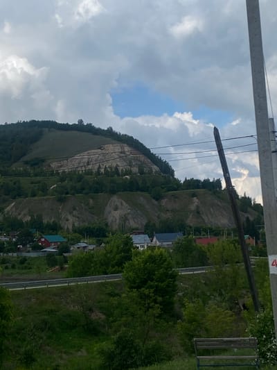 The image depicts a rural landscape in Shiryaevo, Russia, featuring a prominent, terraced hillside with exposed rock formations, suggesting a quarry or natural erosion. Lush green trees and vegetation cover the lower slopes and the foreground, where a small village with several houses and a red-roofed building is visible. A road with a metal guardrail runs horizontally across the middle ground. A utility pole stands on the right side of the frame, with a partially visible red number "4" on it. The sky is partly cloudy, with patches of blue visible amongst the white and grey clouds. The overall scene conveys a sense of natural beauty and rural life.