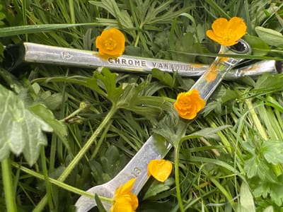 The image depicts two silver-colored wrenches lying in green grass, interspersed with yellow buttercup flowers. The wrenches are labeled "13" and "CHROME VANADIUM". The location is Grange Ville, Ireland. No people are visible. The scene suggests a contrast between industrial tools and natural elements.