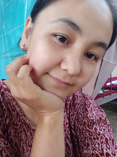 This is a close-up, selfie-style photograph of a young woman. She has dark hair and dark eyes, and her right hand is resting on her chin. She is wearing a red and white patterned top and a gold earring in her right ear. The background is a bit blurry, but it appears to be outdoors, with a blue corrugated metal wall behind her on the left and a bed with a red mattress visible on the right. There is a string or wire hanging from the top right corner of the image. The lighting suggests it is daytime. The timestamp in the bottom right corner reads "30.06.2026 20:05". The woman's expression is neutral.