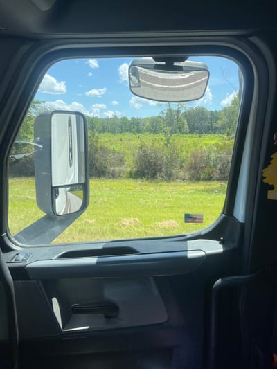 The image is taken from inside a vehicle, likely a truck, looking out the window. The view is of a rural landscape with green grass and trees under a bright blue sky with scattered white clouds. A large side mirror of the truck is prominently featured on the left side of the frame, reflecting the road ahead. A smaller secondary mirror on the side mirror shows a reflection of the road curving to the right. A small American flag sticker is visible on the exterior of the truck door. The interior of the truck is visible in the foreground, including a door handle and part of a seat. The scene appears to be in Riceboro, United States, judging by the context.