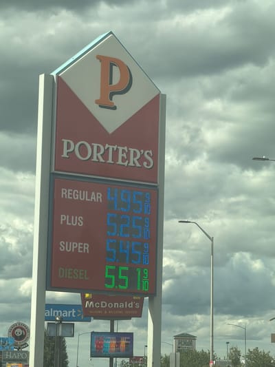 In Pasco, United States of America, a digital gas price sign stands tall against a cloudy sky. The sign displays the name "PORTER'S" prominently at the top, with a large orange "P" logo. Below, the prices for different fuel types are listed: Regular is $4.95, Plus is $5.25, Super is $5.45, and Diesel is $5.51. In the background, other signs are visible, including "Walmart" and "McDonald's," suggesting a commercial area. Streetlights are also present. The overall scene depicts a typical roadside view with commercial establishments under a dramatic, overcast sky.