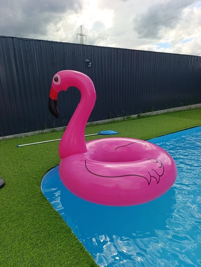 In an outdoor setting, a bright pink inflatable flamingo pool float rests in the blue water of a pool. The pool is bordered by artificial green turf, which is enclosed by a dark gray metal fence. In the background, a utility tower with power lines is visible against a cloudy sky. The scene is devoid of people.
