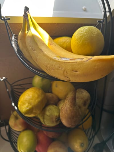 The image shows two black wire fruit baskets. The basket on the left contains bananas and lemons. The basket on the right contains pears, lemons, apples, and a lime. The location of the image is unknown and is not stated. There are no visible people, actions, or interactions in the image.