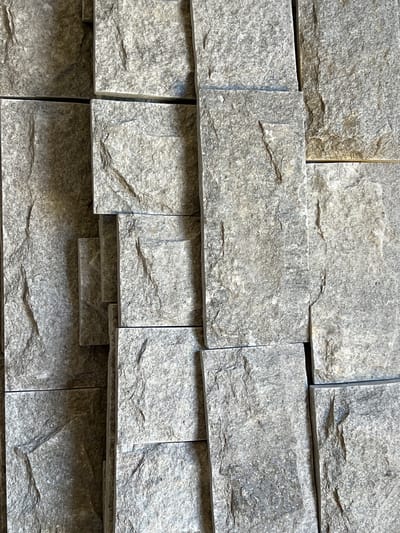 The image shows a close-up view of a wall constructed from rough, grey stone tiles. The tiles are rectangular and vary slightly in size and shape, creating an uneven, textured surface. The stone has a natural, irregular grain with visible cracks and fissures, giving it a rustic appearance. The lighting suggests an outdoor setting, with shadows cast across the tiles, highlighting their three-dimensional texture. There are no people or discernible objects other than the stone wall itself. The scene is devoid of any specific indicators of its location within Menenkyovo, Bulgaria, as the image focuses solely on the architectural detail of the wall.
