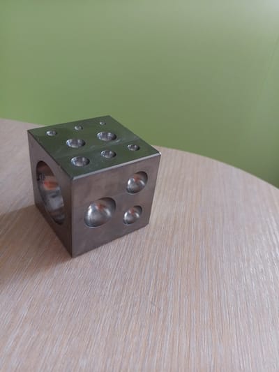 A close-up, eye-level, medium shot captures a metallic cube with multiple hemispherical indentations on its surfaces, resting on a light-colored wooden table. The cube is positioned on the left side of the frame, with its top and side surfaces visible. The indentations vary in size and depth. The background features a solid, light green wall. The scene is indoors, with a light source casting reflections on the metallic surface of the cube and the wooden table. The image does not provide any information that could place its location in Vilyuysk, Russia, or any other specific city. There are no people, actions, or interactions depicted.