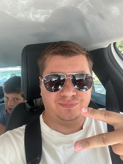 The media file captures a selfie taken by a man in a car. He is wearing aviator sunglasses and a white t-shirt, and he is holding up his hand in a peace sign. The man appears to be smiling, and the overall mood is casual and lighthearted.
The background of the image shows the interior of a car, with a headrest and a seatbelt visible. There is also a glimpse of another person, a young boy, in the backseat. The boy is looking directly at the camera with a curious expression. The scene is lit by natural daylight, suggesting it is daytime. The reflection in the man's sunglasses shows trees and buildings, indicating an outdoor setting. The location context provided is Benimàmet-beniferri, Spain. There is no visible text in the image.