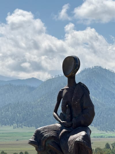 A dark bronze sculpture of a seated, faceless human figure is positioned in the foreground against a backdrop of layered, forested mountains and a bright blue sky with scattered white clouds. The sculpture depicts a figure with its head tilted back and its face an empty oval opening. The figure is draped in fabric that flows around its legs and shoulders, and its arms are held in front, cradling something small. The sculpture is weathered and appears to be made of metal, with visible patina and texture. The surrounding environment suggests an outdoor setting, likely a park or scenic overlook, with a valley visible in the distance between the sculpture and the mountains. The location is identified as Bichiktu-Boom, Russia.
