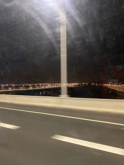 The photo displays an elevated roadway at night, viewed through a window. The road, marked with white lines, dominates the right side. On the left, a blurred expanse reveals distant city lights reflected in water, creating a sparkling effect. The window's surface shows light streaks and reflections, adding to the atmospheric quality.