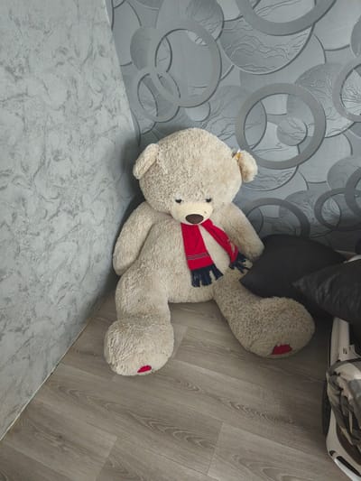 An oversized, light beige teddy bear sits on a light brown wood-grain floor in the corner of a room. The bear is wearing a red and blue scarf around its neck. The wall behind the bear has a textured grey pattern. To the right of the bear, two dark grey pillows are visible, along with a portion of a white object with a folded grey and white checkered blanket on top, possibly a stroller or a bed. The room appears to be indoors, with no external environmental features visible.