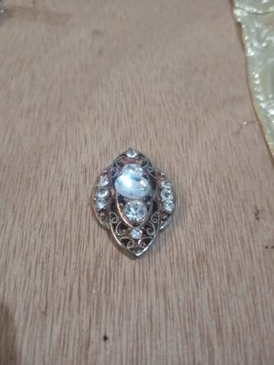 A decorative, almond-shaped embellishment is centrally positioned on a light brown wooden surface. The object measures approximately 3-4 cm in length, featuring a metallic setting with intricate scrollwork and multiple clear, faceted stones. Two prominent oval-cut stones are vertically stacked in the center; the upper stone is slightly larger than the lower one. Smaller round-cut stones encircle the central stones and are integrated into the filigree detailing. The wooden surface displays visible grain patterns and minor imperfections. A blurry, golden, textured fabric is partially visible in the upper right quadrant of the frame. The scene is located in Chandragiri, Nepal.